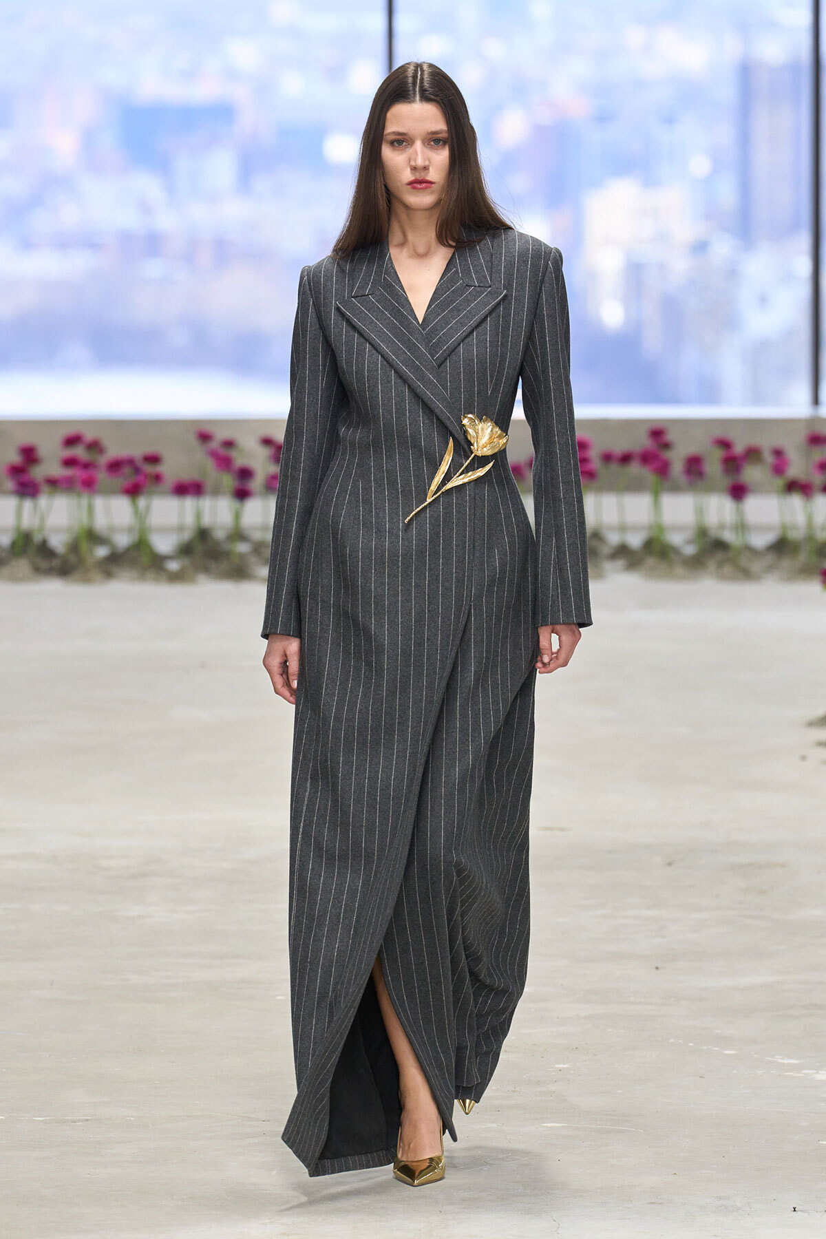 Model wearing a dark gray pinstripe suit with a gold floral brooch at the waist, walking on a runway.
