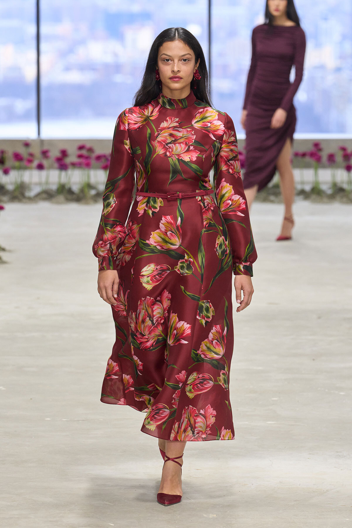 Model walking a runway in a burgundy floral satin midi dress with a thin belt and red heels.