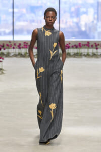 Female model walks a pinstripe gray jumpsuit with gold floral embroidery on a runway indoors.