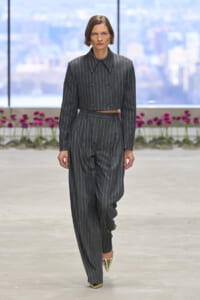 Model in a dark gray pinstripe suit with a cropped jacket and wide-leg pants walking a runway; gold pointed heels, city skyline in the background.