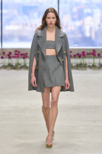 Model walks the runway in a gray two-piece set: a cape-style coat over a crop top and matching mini skirt, with gold heels.