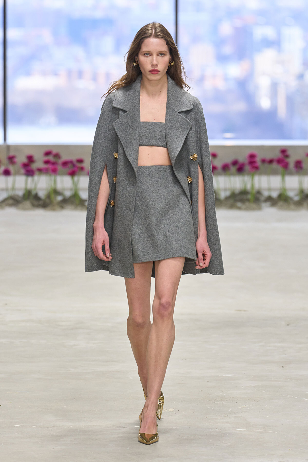 Model walks the runway in a gray two-piece set: a cape-style coat over a crop top and matching mini skirt, with gold heels.