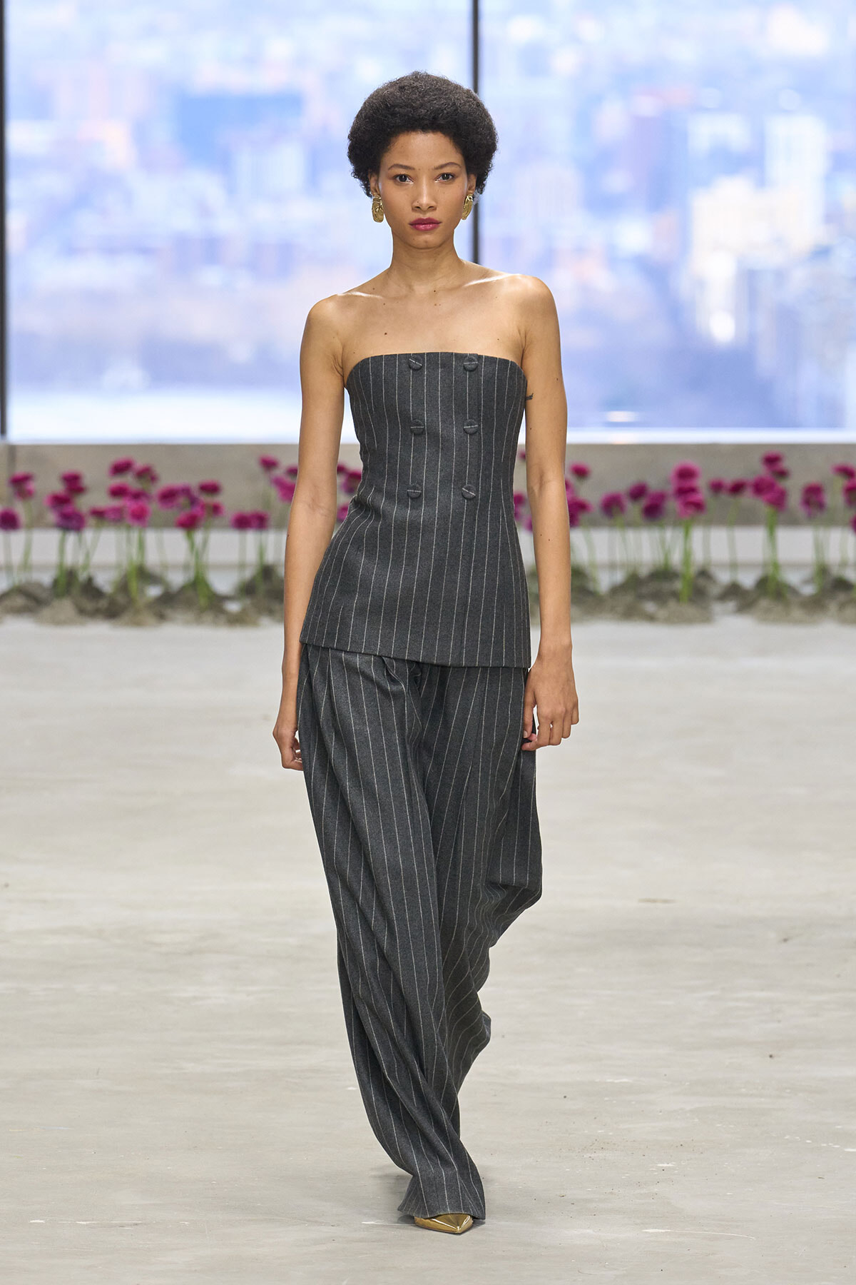 Model walking a fashion runway in a dark pinstripe strapless top and wide-leg pants, cityscape backdrop behind floral planter beds.