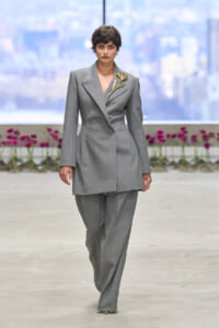 Model walks the runway in a tailored gray pantsuit with a gold leaf brooch on the left shoulder, against a cityscape backdrop with pink flowers in the foreground.