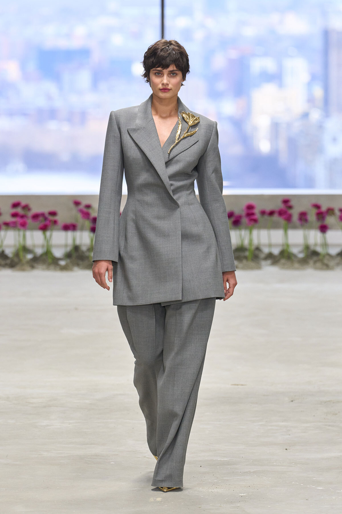 Model walks the runway in a tailored gray pantsuit with a gold leaf brooch on the left shoulder, against a cityscape backdrop with pink flowers in the foreground.