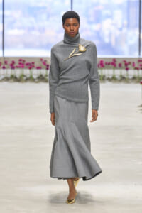 Model walking a grey turtleneck sweater with a gold flower brooch and flowing grey skirt on a runway, gold pointed heels visible.