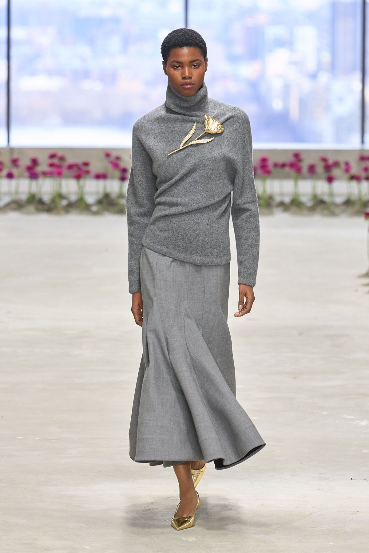 Model walking a grey turtleneck sweater with a gold flower brooch and flowing grey skirt on a runway, gold pointed heels visible.