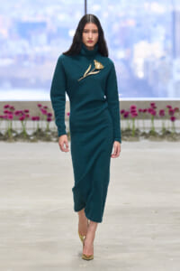 Model walking a teal turtleneck dress with a gold flower brooch on the chest, gold pointed heels, runway setting with pink flowers in the background.
