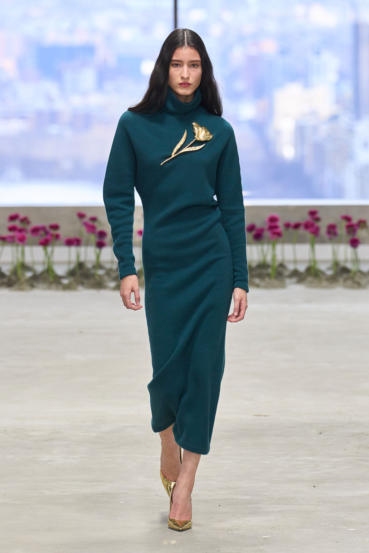 Model walking a teal turtleneck dress with a gold flower brooch on the chest, gold pointed heels, runway setting with pink flowers in the background.