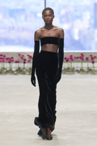 Model on a runway wearing a black velvet off-the-shoulder bandeau top, long gloves, and a floor-length velvet skirt.