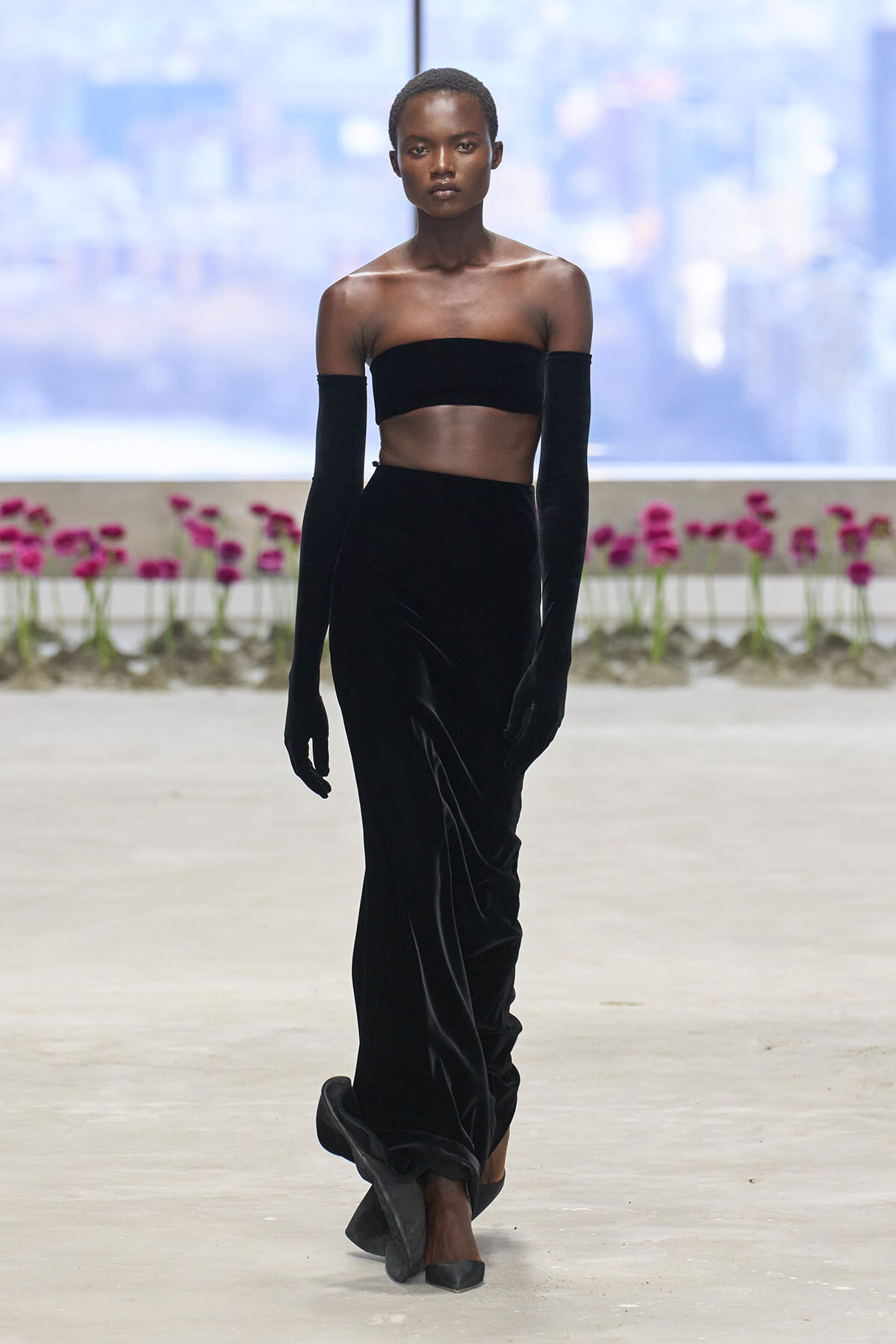 Model on a runway wearing a black velvet off-the-shoulder bandeau top, long gloves, and a floor-length velvet skirt.