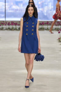 Model on a runway wearing a cobalt blue sleeveless dress with two vertical rows of gold buttons, carrying a matching blue handbag and blue heels.