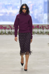 Model walking a runway in a deep plum turtleneck sweater and a knee-length purple lattice-knit skirt adorned with silver beads, paired with black pointed heels and oversized black sunglasses.