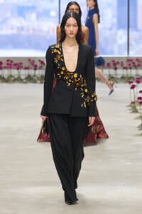 Model walking a runway in a black blazer with deep plunge and gold floral embroidery along the bodice.