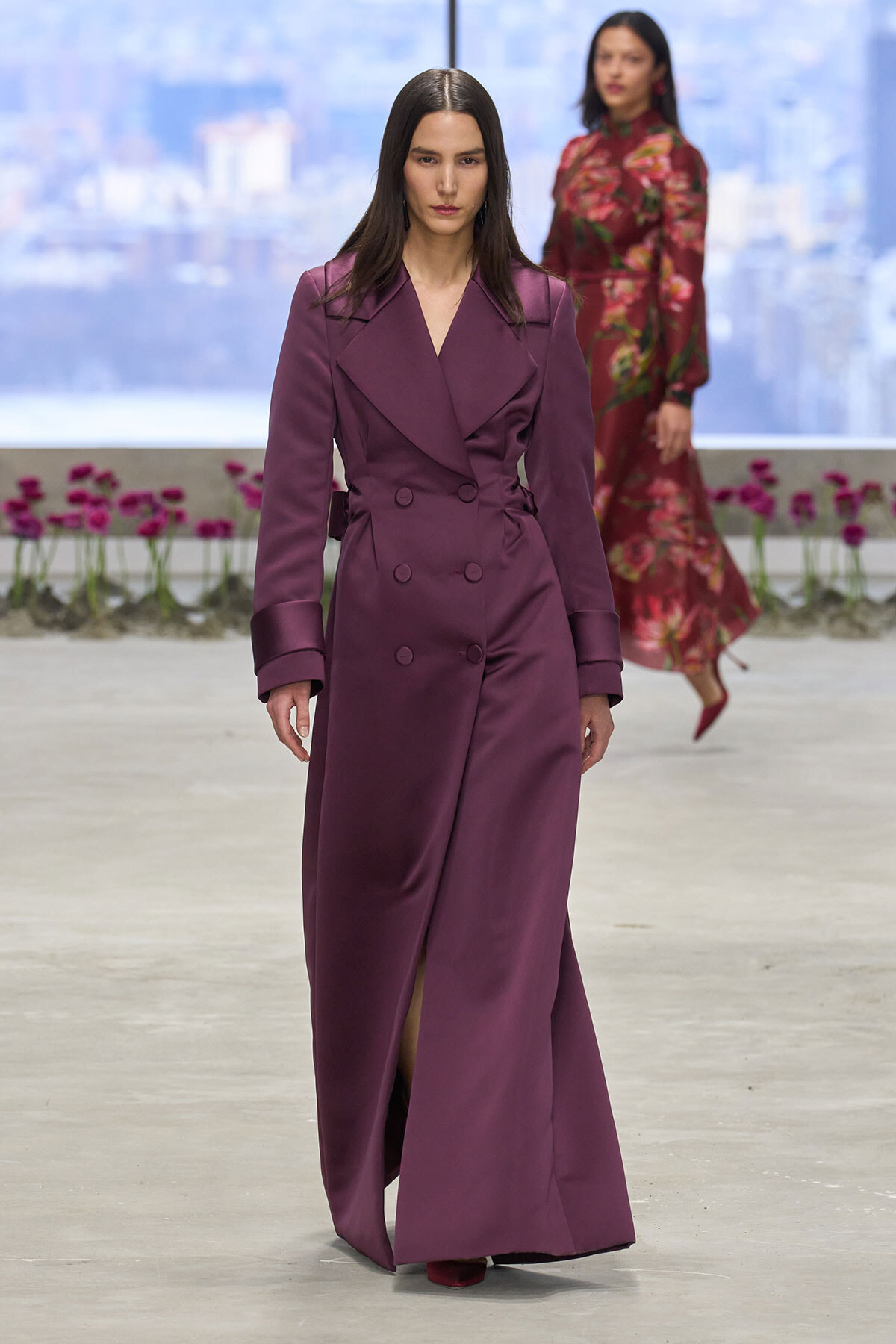 Model walking the runway in a long plum double-breasted trench coat with oversized lapels and matching belt.