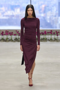 Model walking a runway in a burgundy, ruched long-sleeve midi dress with a draped skirt and matching burgundy heels on a neutral catwalk backdrop.