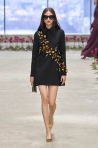 Model walking the runway in a black mini dress with gold floral embroidery, gold heels, and oversized sunglasses, holding a small clutch.