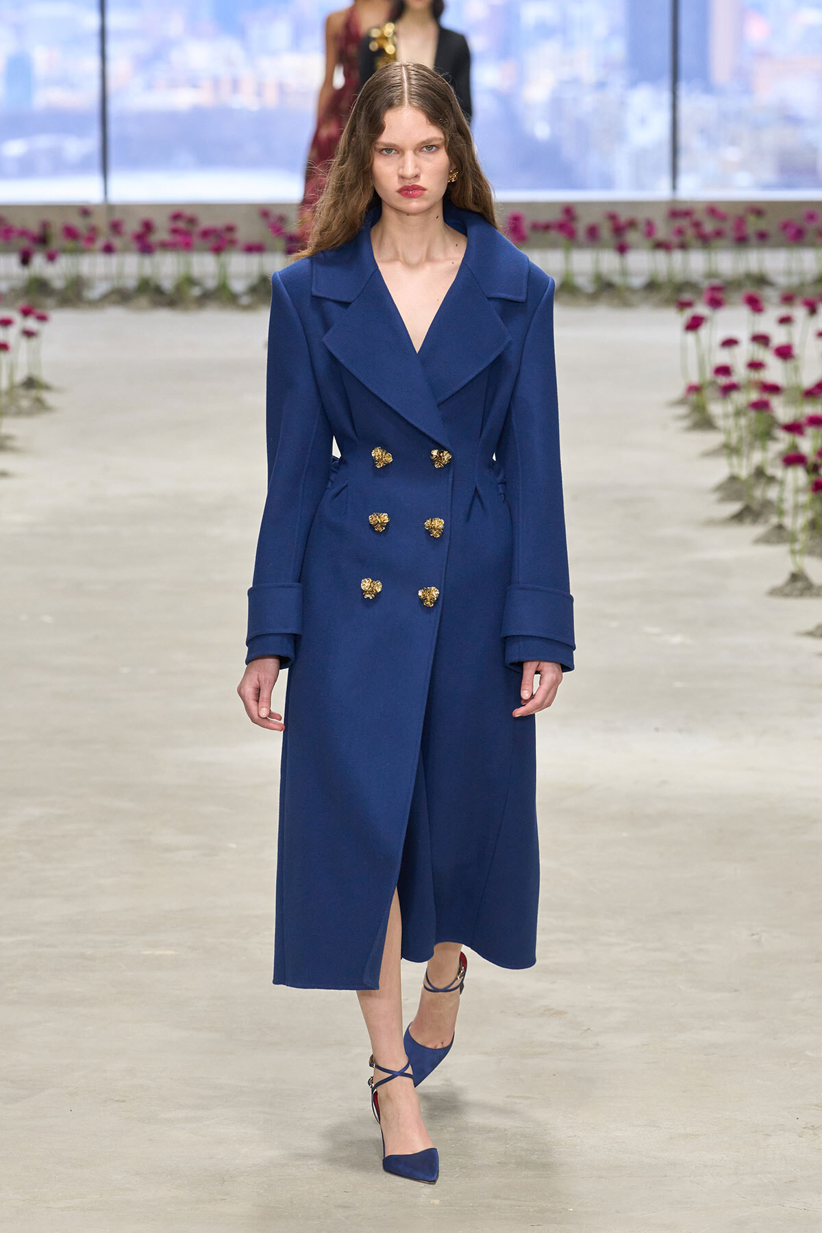 Model walking a runway in a royal blue double-breasted coat with gold buttons and matching blue heels.