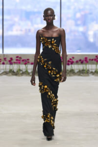 Model walking a runway in a black strapless gown with gold floral embroidery cascading from the bodice to the skirt.