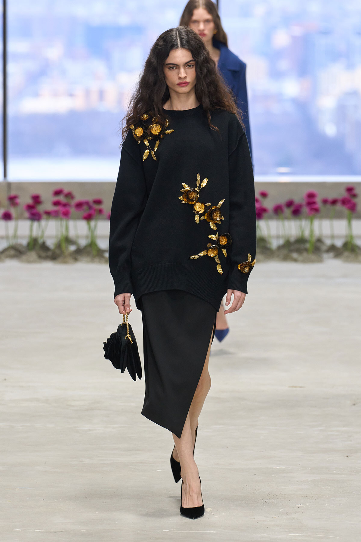 Model walking a runway in a black knit sweater with gold floral appliqués and an asymmetric black skirt, holding a small black clutch with a gold chain.