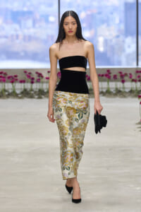 Model walking a runway in a black bandeau top with a cutout, metallic floral trousers, black heels, and a small black clutch against an urban backdrop.