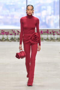 Runway model in a burgundy turtleneck jumpsuit with a large fabric rose at the hip, carrying a matching rose-adorned handbag on a minimalist catwalk.