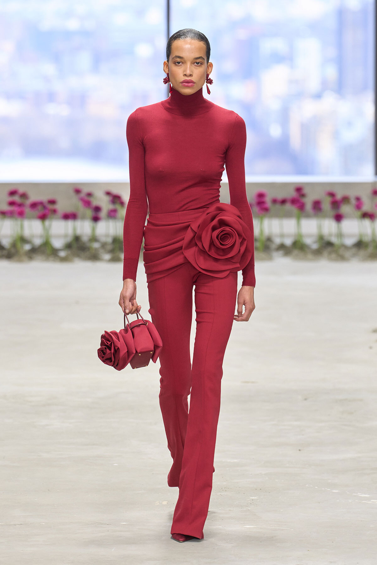 Runway model in a burgundy turtleneck jumpsuit with a large fabric rose at the hip, carrying a matching rose-adorned handbag on a minimalist catwalk.