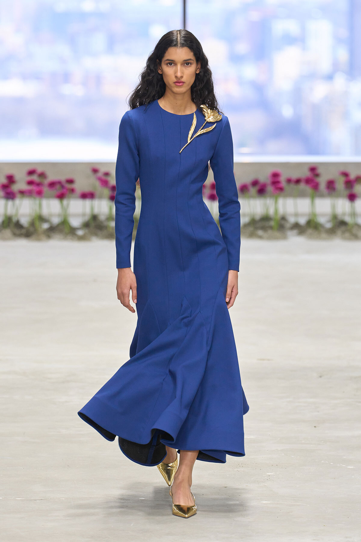 Model walks a royal blue, long-sleeve gown with gold leaf brooch, gold pointed heels, in an urban backdrop with pink flowers lining a terrace.