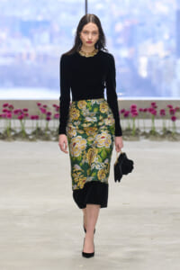 Model walking runway in a black long-sleeve top and green floral midi skirt with gold embroidery, wearing gold necklace and black heels.