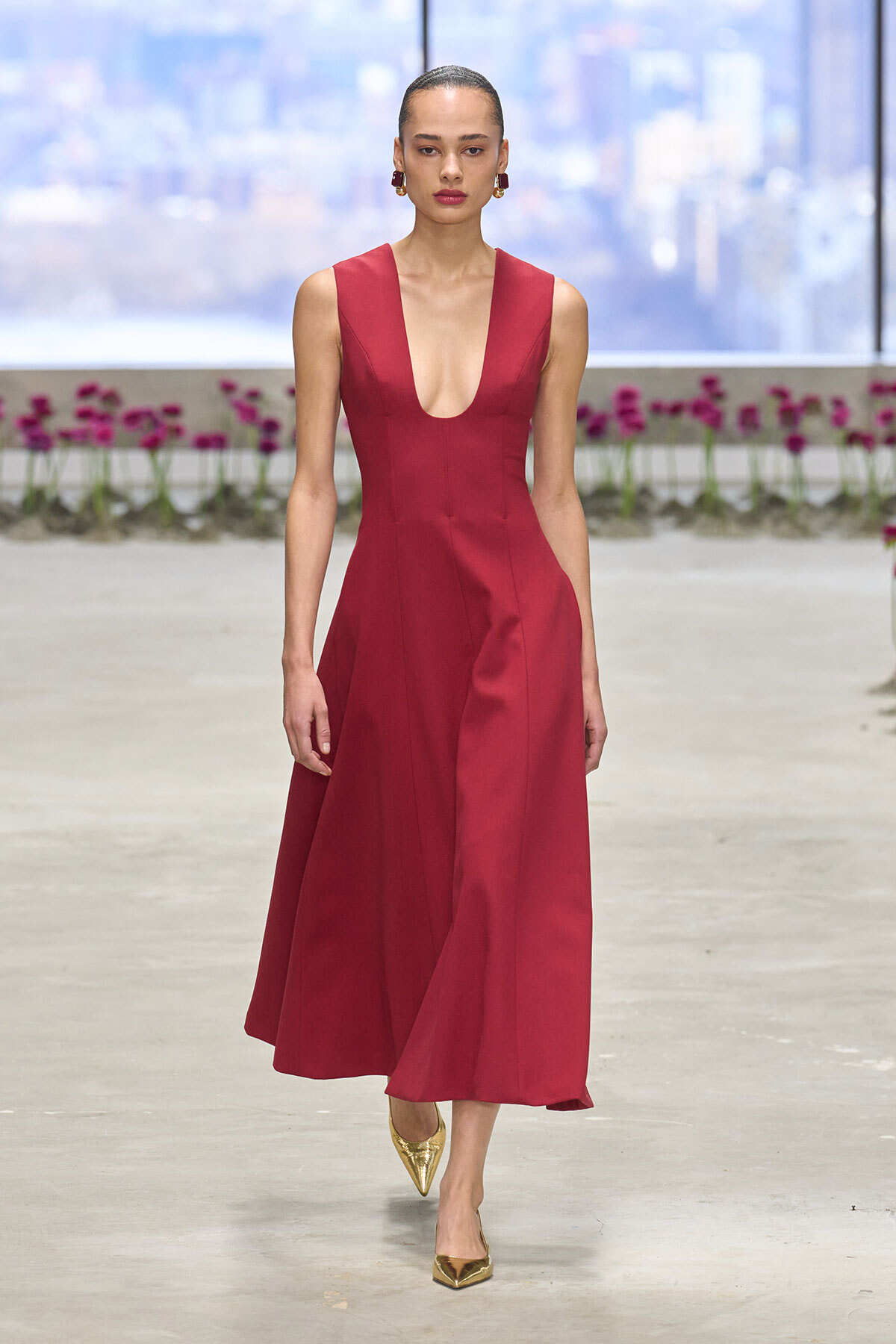 Model walking a runway in a sleeveless red gown with a deep V neckline and flared skirt, wearing gold pointed heels.