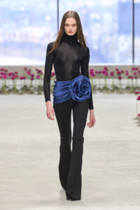 Model on a fashion runway wearing a black sheer turtleneck and fitted black pants with a royal blue satin waist sash forming a large rose.