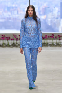 Model walking a runway in a light blue floral lace pantsuit with a high-neck top, against a city backdrop.