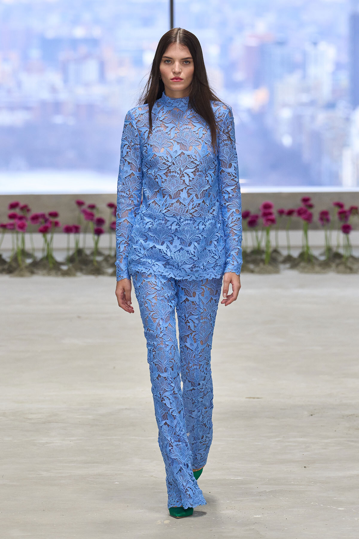 Model walking a runway in a light blue floral lace pantsuit with a high-neck top, against a city backdrop.