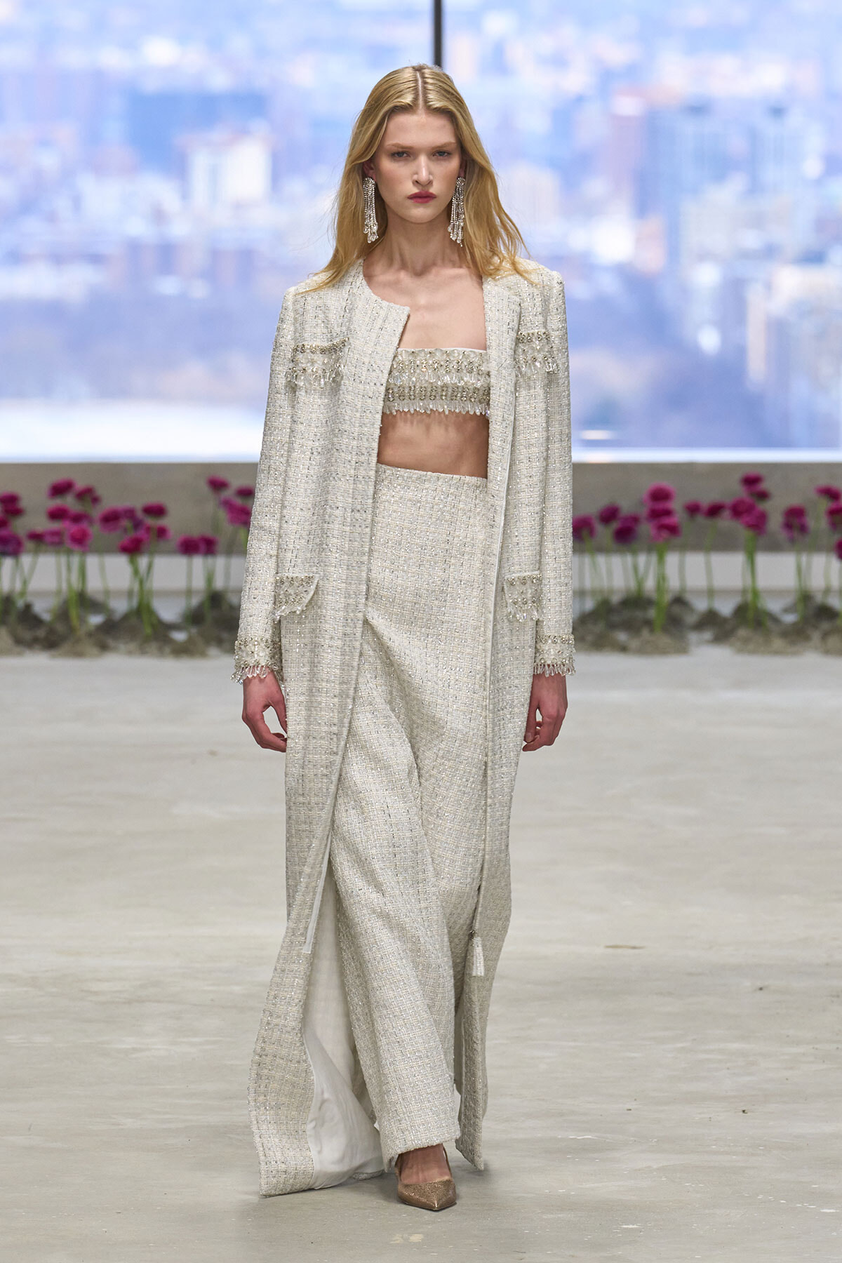 Model walking a runway in a cream tweed matching suit with a long coat, fringed crop top, and wide-leg trousers; statement chandelier earrings and nude heels.