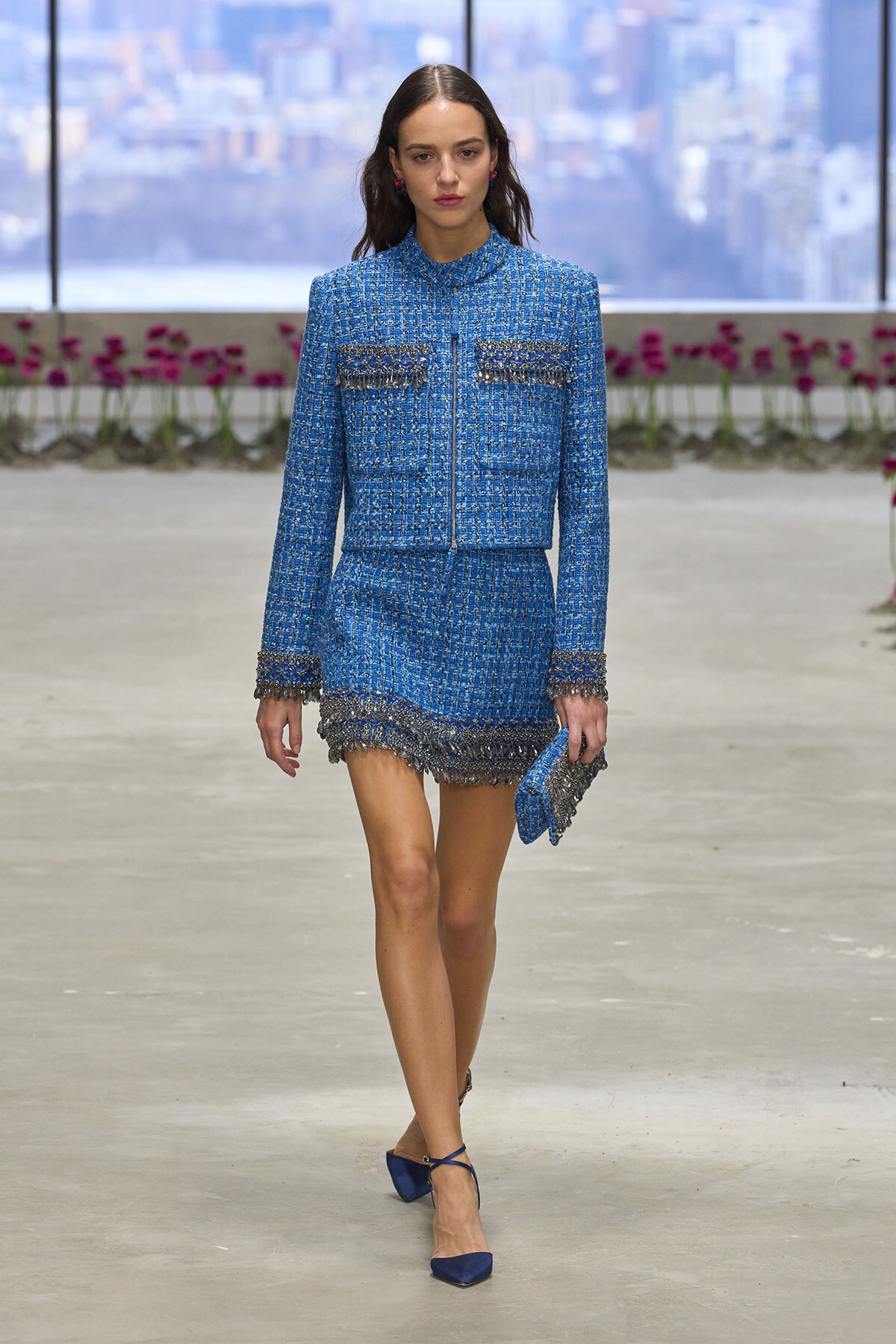 Model walks a runway in a blue tweed fringe-trimmed suit—jacket and mini skirt—carrying a matching blue clutch and wearing blue heels, with a blurred cityscape in the background.