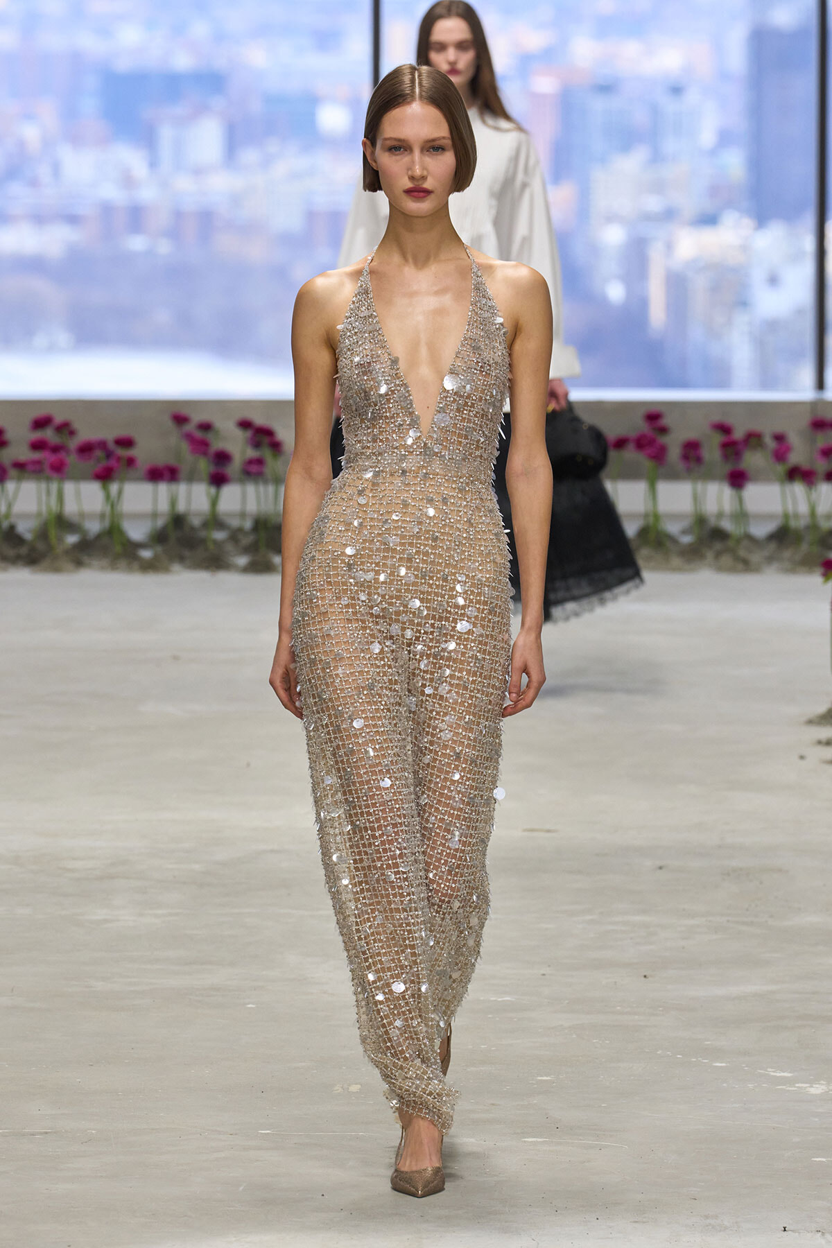 Model walks runway in a silver beaded halter jumpsuit with a plunging neckline and wide legs, sparkling under lights against a city backdrop