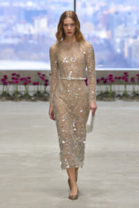 Model on a runway wearing a sheer, beaded champagne dress with a thin belt, holding a matching beaded clutch.
