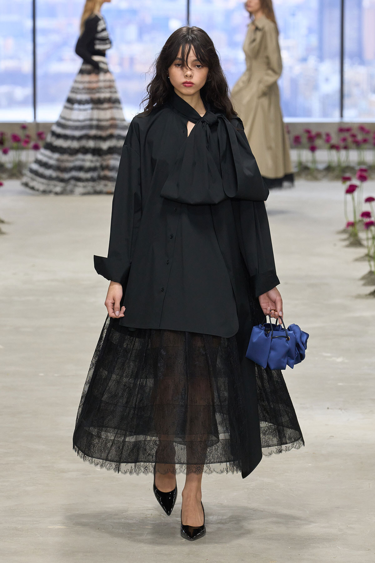 Model walking the runway in a black bow-tossed blouse and a sheer black lace midi skirt, carrying a blue handbag.