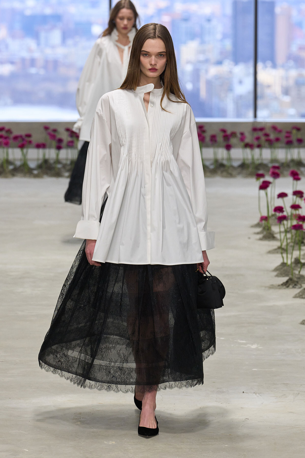 Model walks a runway in a white pleated-front blouse and black lace midi skirt, carrying a small black handbag, with pink flowers and cityscape in the background.