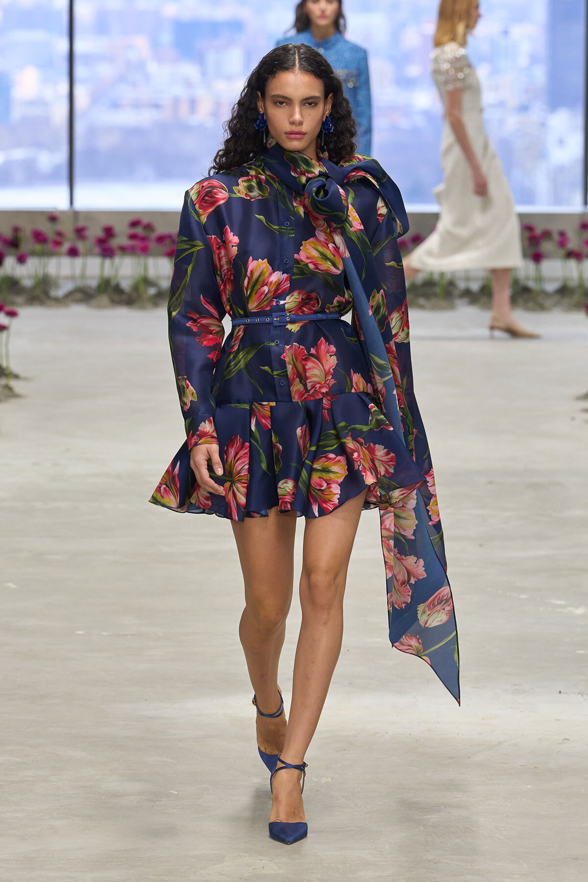 Model walking a fashion runway in a navy floral print dress with a tied scarf and blue heels.