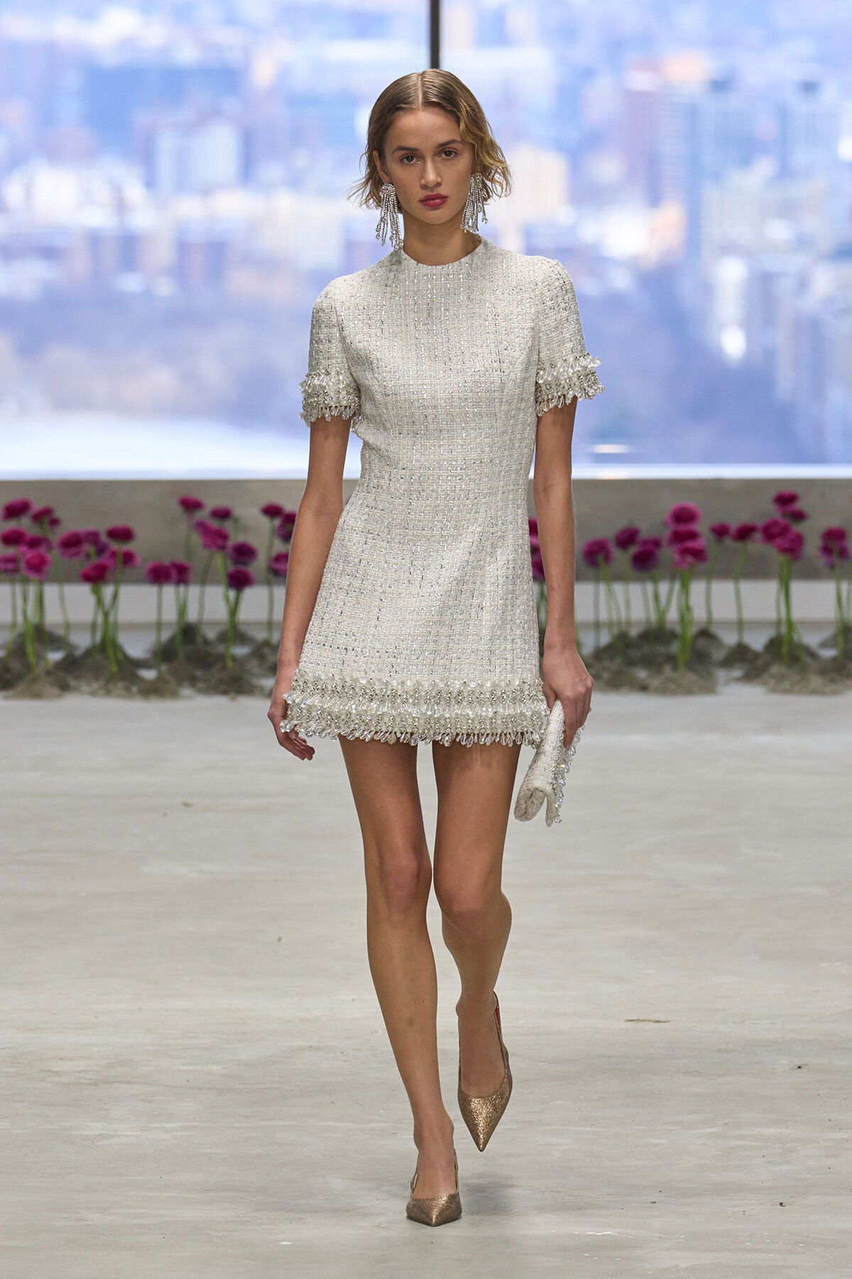 Model walking a runway in a silver tweed mini dress with fringe trim on sleeves and hem, city skyline in background.
