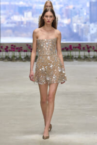 Model walks runway in a strapless beige mini dress covered in silver sequins and circles, paired with metallic flats, urban backdrop behind.
