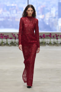 Model walking a runway in a burgundy lace high-neck gown with long sleeves and sheer panels, city skyline backdrop behind.