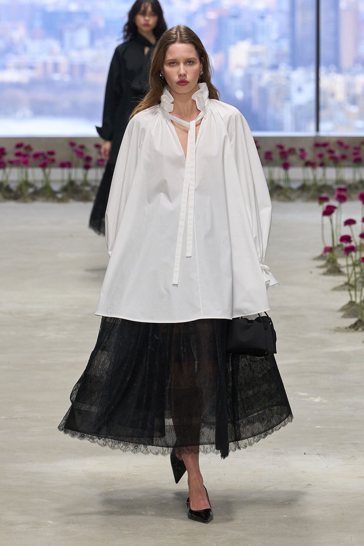 Model walks a fashion runway in a white voluminous blouse over a black lace skirt, carrying a small black bag.