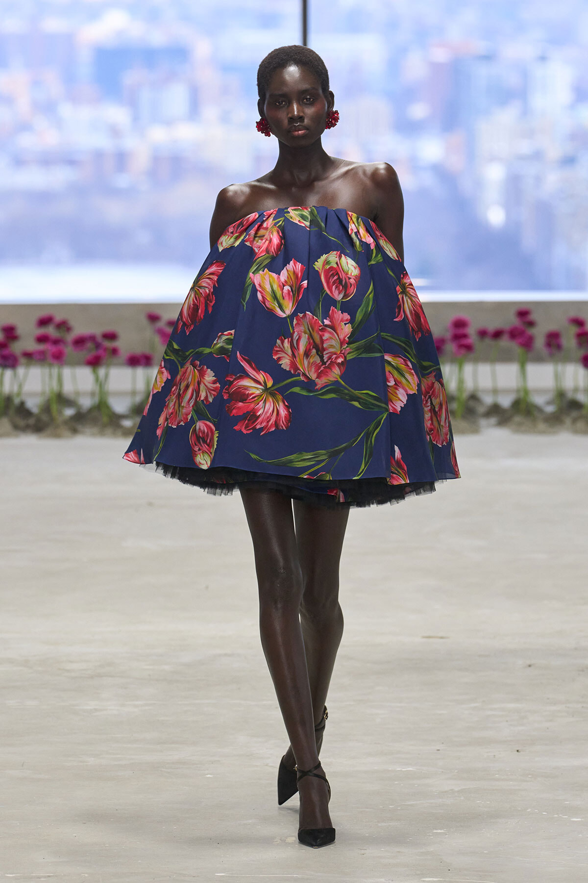 Model walking a fashion runway in an off-shoulder navy dress with large pink flowers, red earrings, and black heels, city backdrop behind.