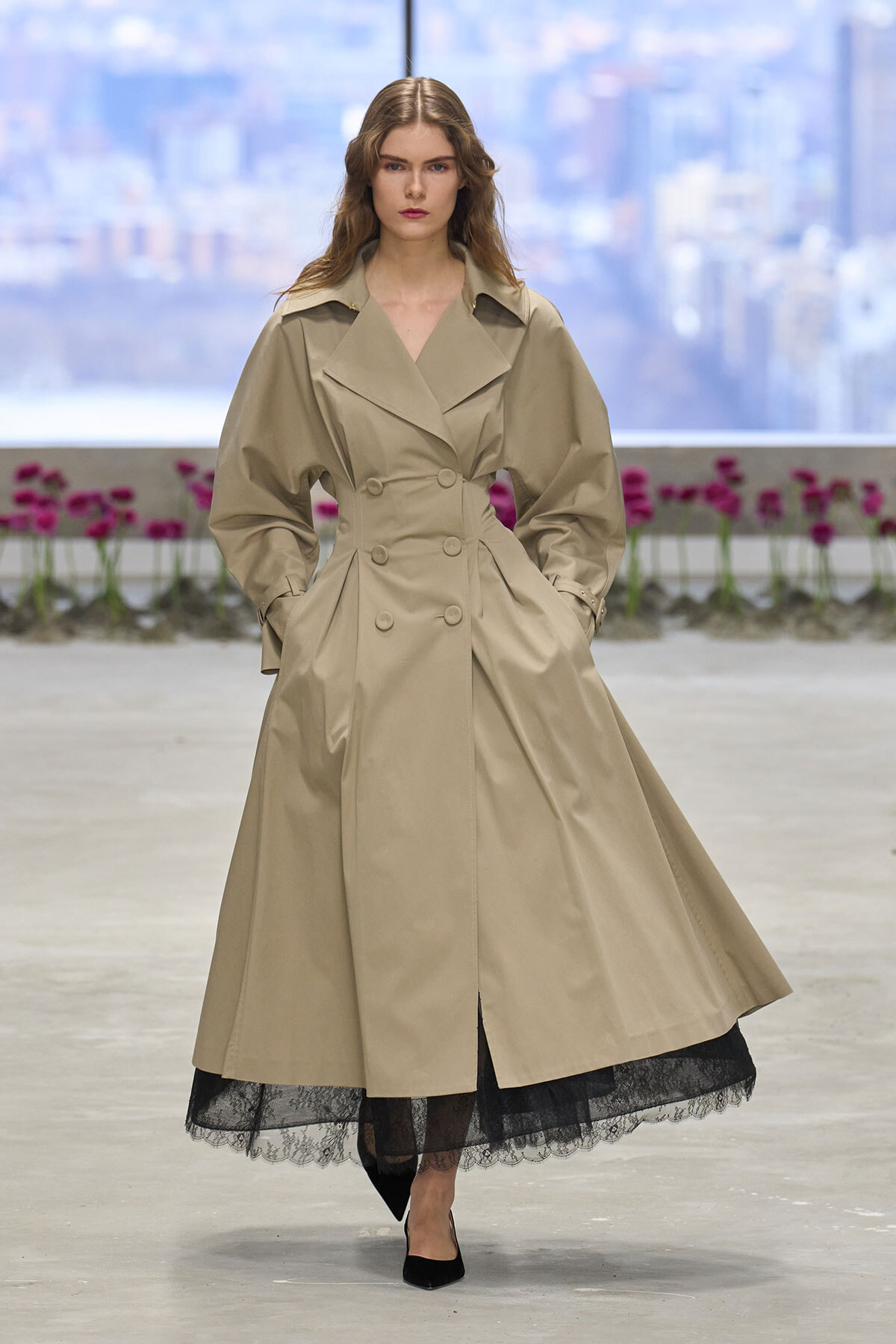 Model walks the runway in a long beige trench coat with a black lace hem showing underneath.