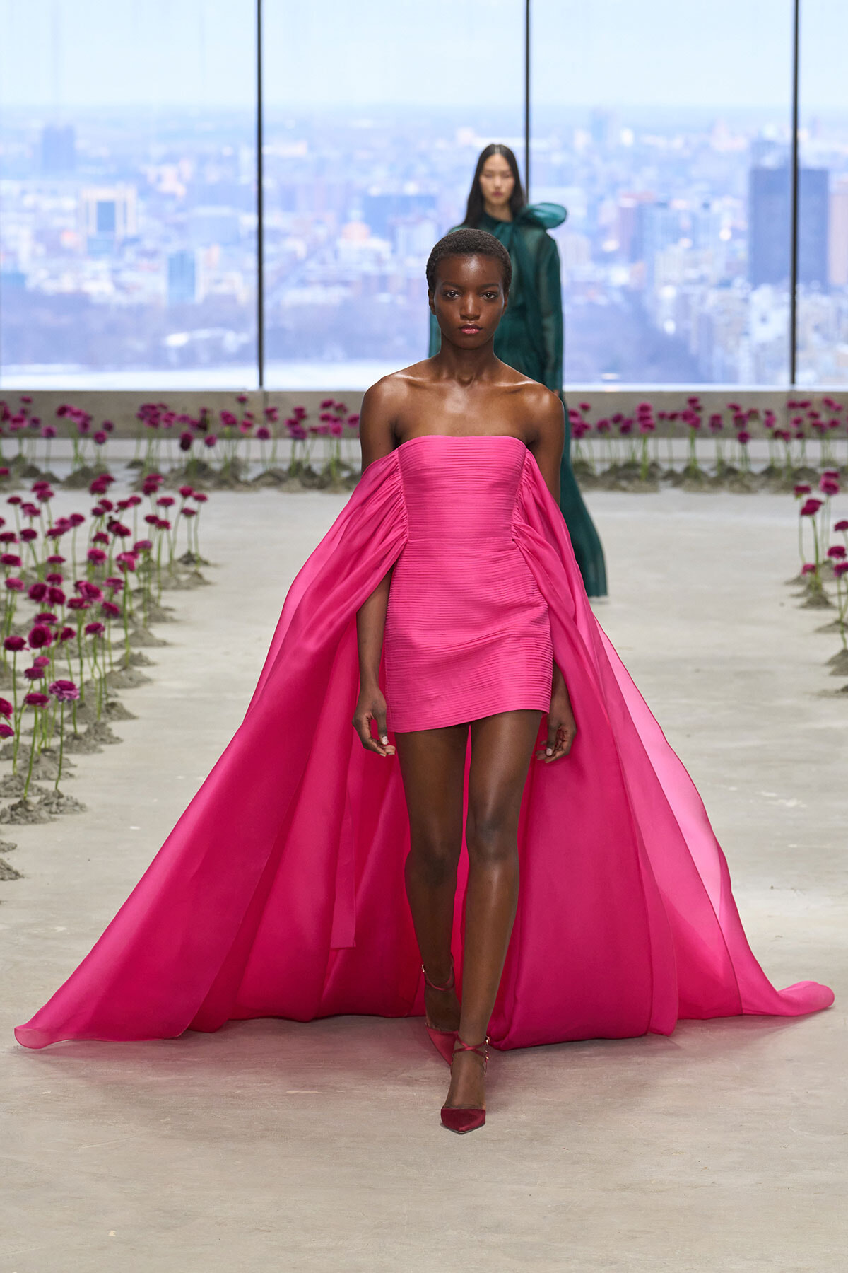 Model walks a fashion runway in a bright pink off-the-shoulder mini dress with a long matching cape flowing behind her, city skyline in the background.