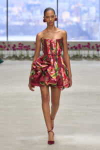 Model walks the runway in a strapless red floral dress with a large sculptural ruffle at the hip, paired with red heels.