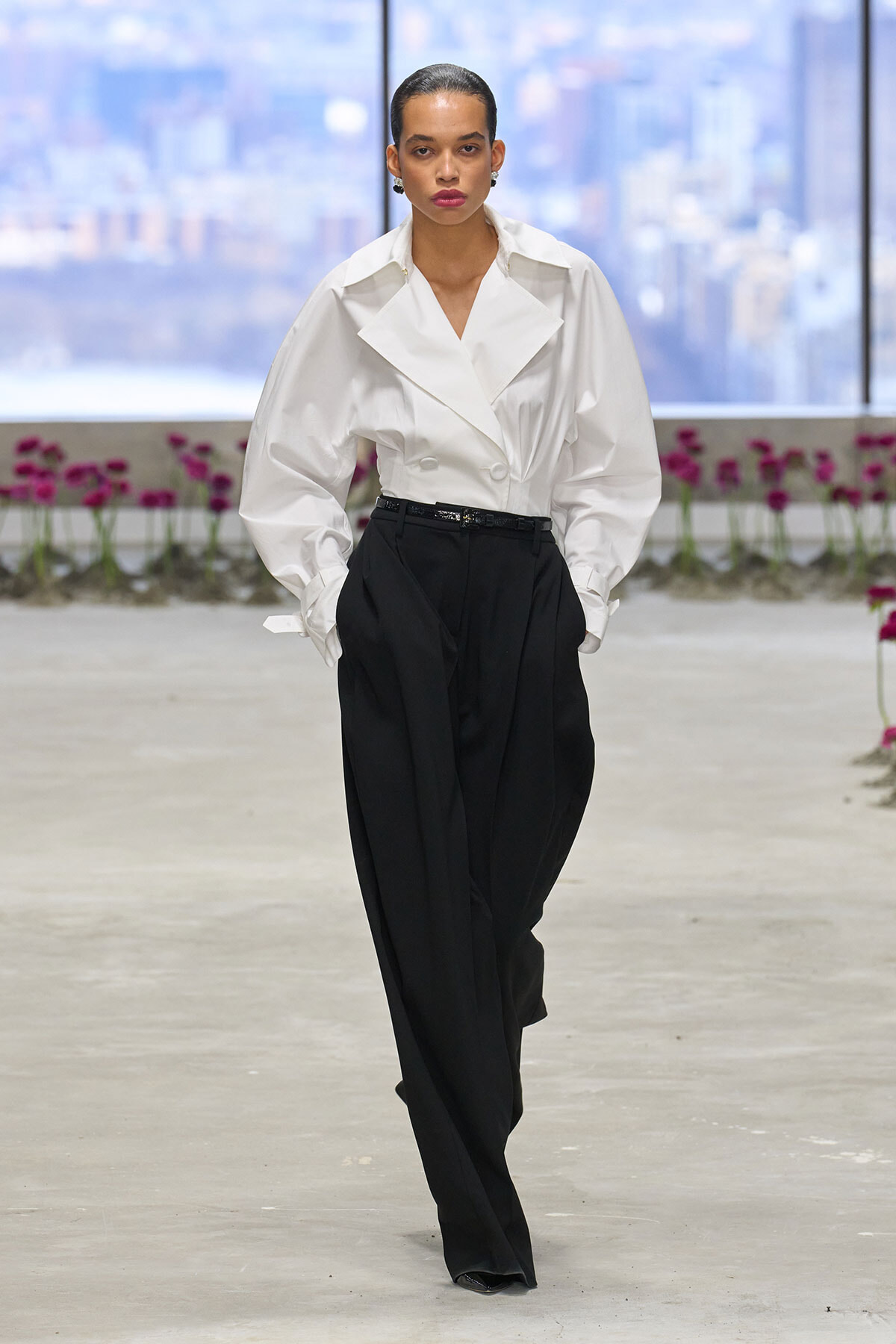 Model walking a runway in a white oversized blouse and black wide-leg trousers, with a citylike backdrop and pink flowers in the background.