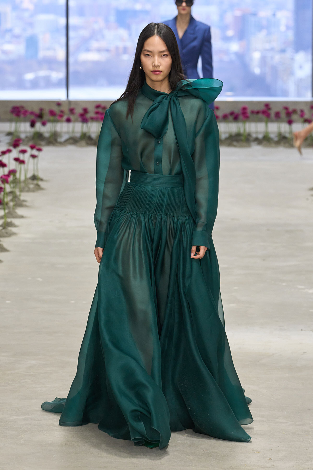 Model walking a runway in an emerald green sheer blouse with a large bow at the neck and a high-waisted, pleated floor-length skirt; cityscape backdrop visible behind.